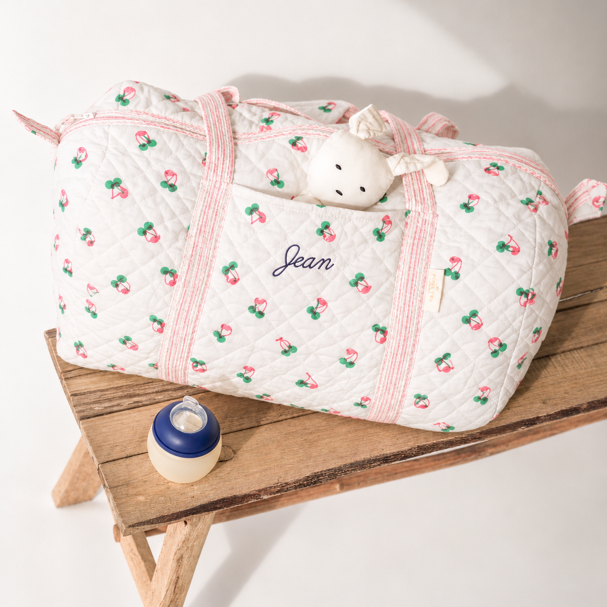 Sac de week-end matelassé à motif cerises avec prénom brodé sur la poche frontale, posé sur un banc en bois